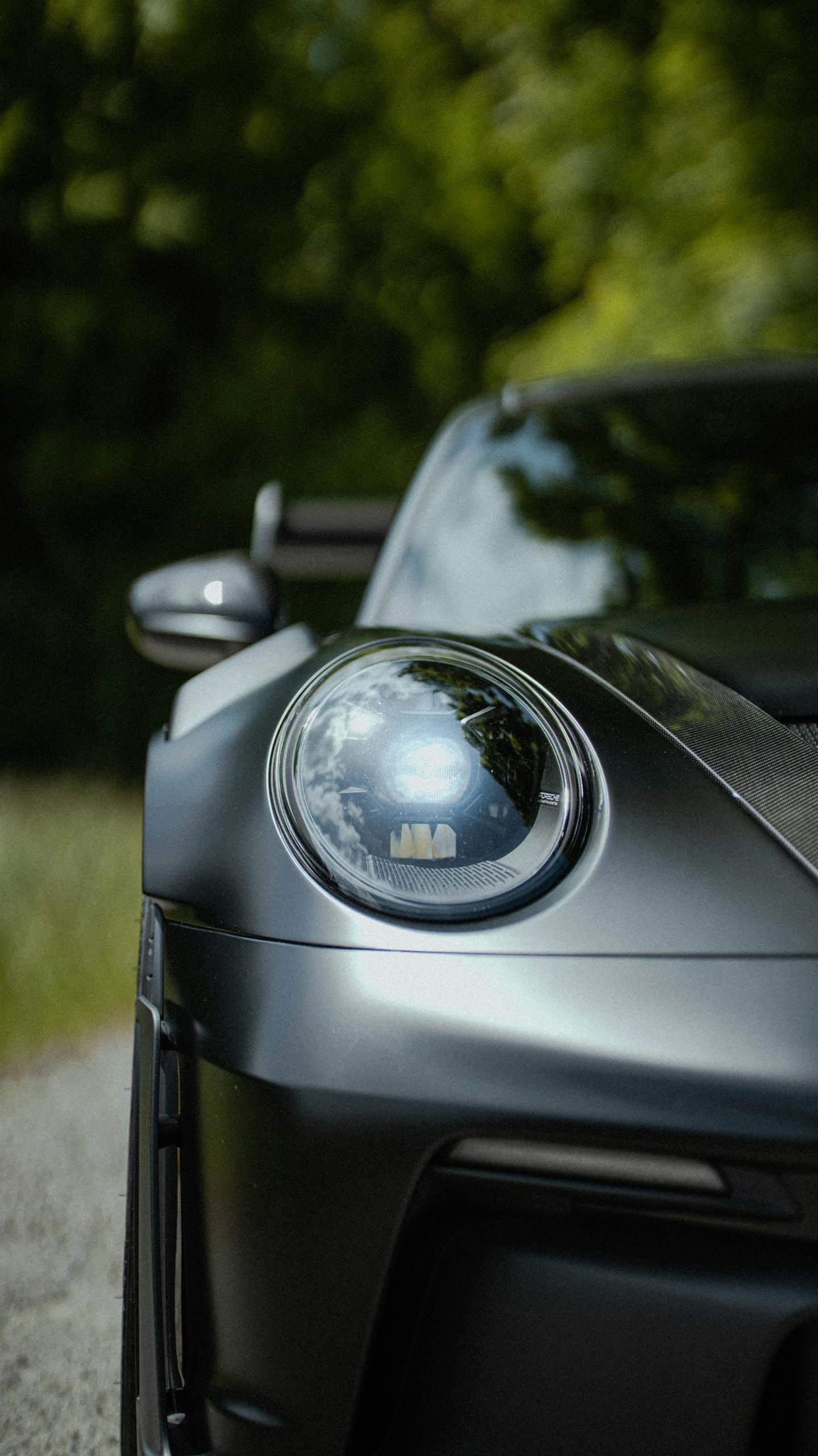 Close-up of a sleek sports car headlight in a forest setting, showcasing elegance and speed.